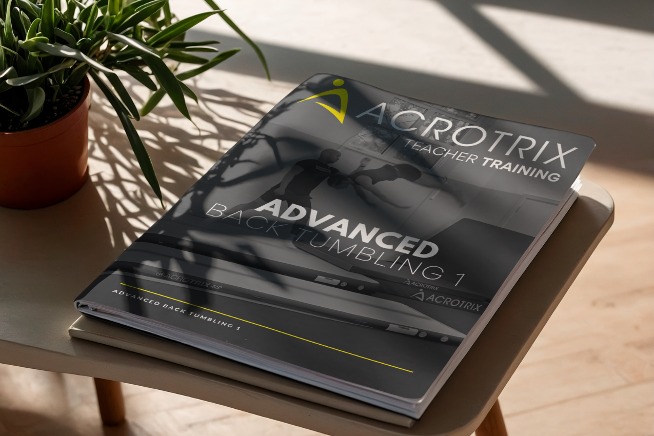 A printed training manual titled 'ACROTRIX TEACHER TRAINING ADVANCED BACK TUMBLING 1' rests on a small wooden table beside a potted plant in a sunlit indoor setting.